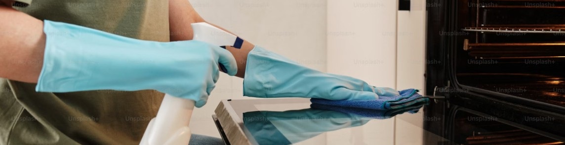 Person med blå handskar rengör glaset på en öppen ugnslucka med trasa och sprayflaska